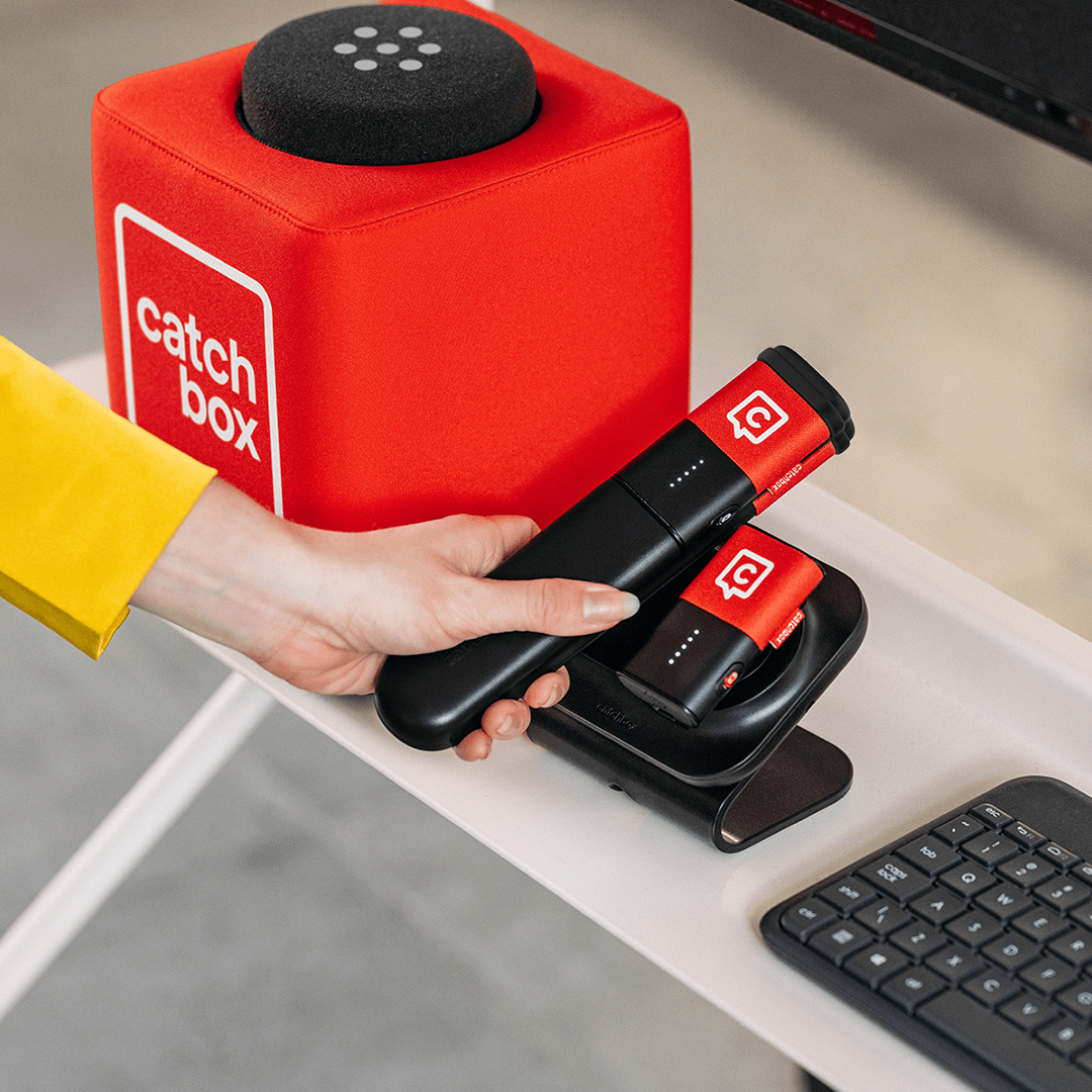 A person's hand reaching for a Catchbox Stick microphone, placed next to a Catchbox Cube and Clip microphone.