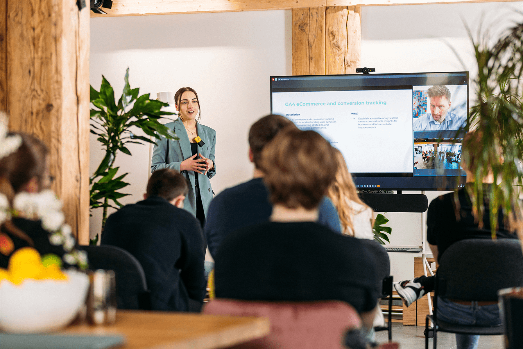 Hybrid meeting with on-site attendees, featuring a presentation leader standing next to a screen, wearing a Catchbox Clip wearable microphone with a lanyard for hands-free presentation and voice amplification.