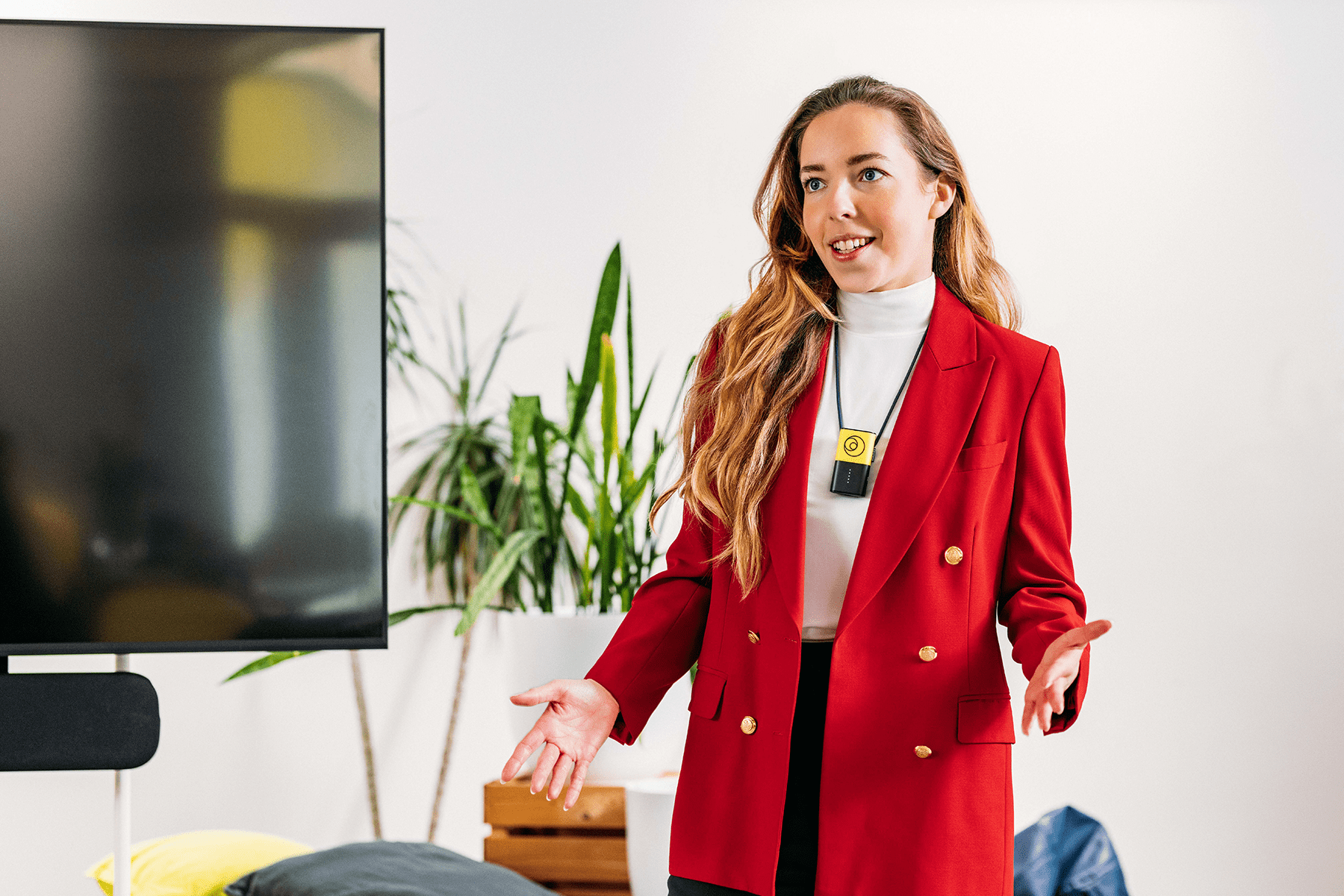 Hybrid meeting with on-site attendees, featuring a presentation leader standing next to a screen, wearing a Catchbox Clip wearable microphone with a lanyard for hands-free presentation and voice amplification. The meeting is being recorded for future reference.