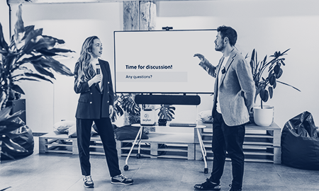 Two presenters stand in front of a mobile cart with a TV screen, amplifying sound using Catchbox Clip and Stick microphones.