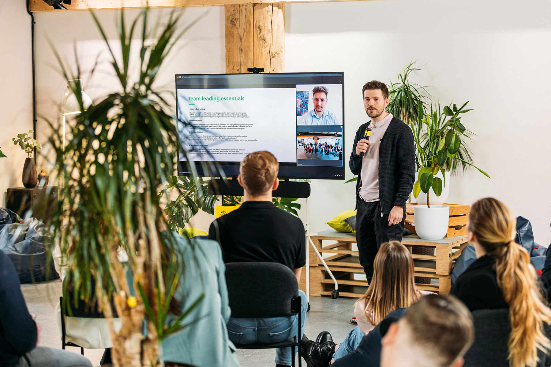 Presenter standing in front of a mobile cart with a TV screen, amplifying sound for audience by capturing audio with Catchbox Stick microphone.